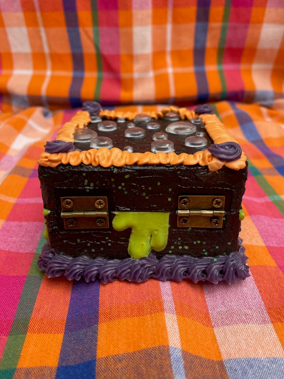 a small hinged box painted black and decorated to look like a cake with black, purple, and orange icing and with green drips on the side
