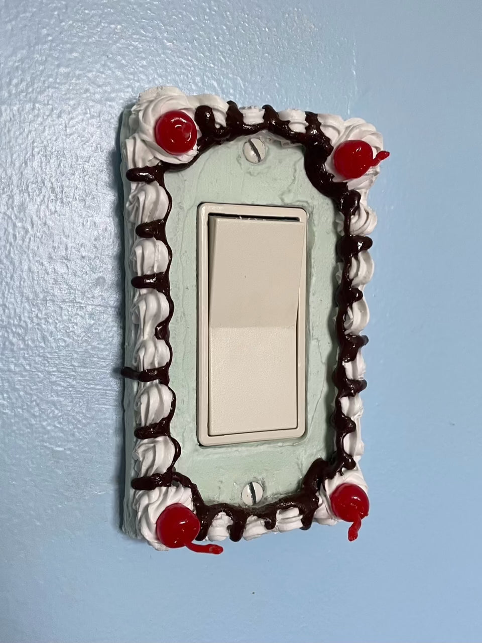 a light switch with a light switch plate decorated to look like a mint green cake with fake cherries and chocolate drizzle