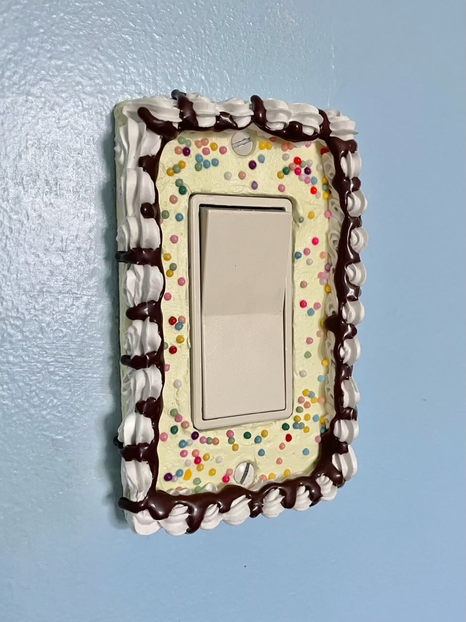 a light switch with a light switch plate decorated to look like a vanilla cake with sprinkles and chocolate drizzle