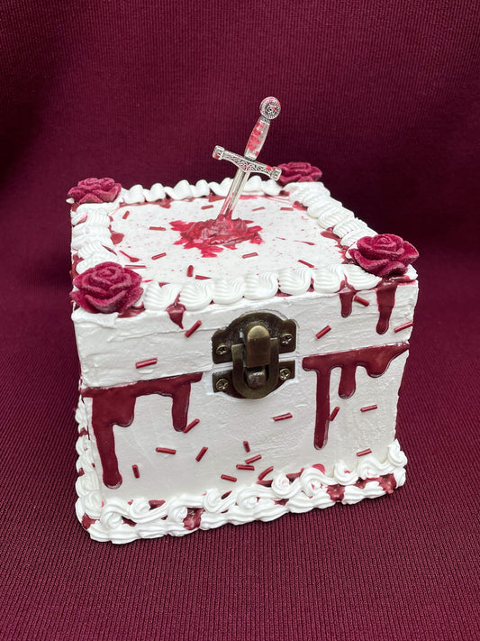a hinged box decorated to look like a white cake with imitation blood, burgundy sprinkles and a mini sword