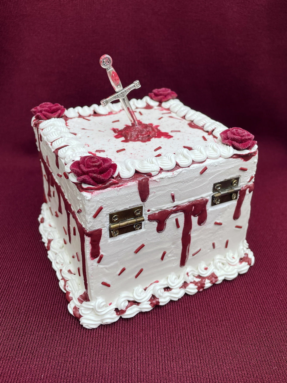 a hinged box decorated to look like a white cake with imitation blood, burgundy sprinkles and a mini sword