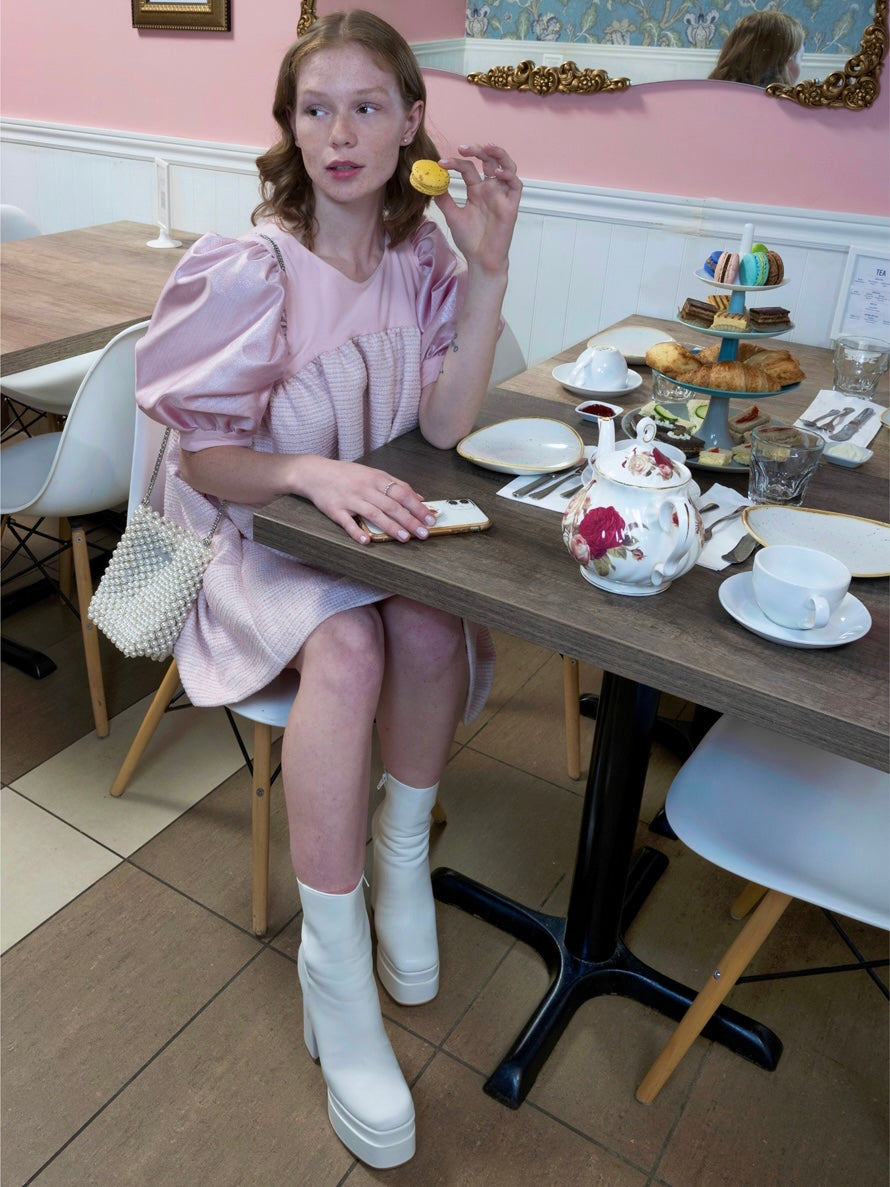 a model wearing a baby pink dress, sitting at a table set with a high tea and pastries