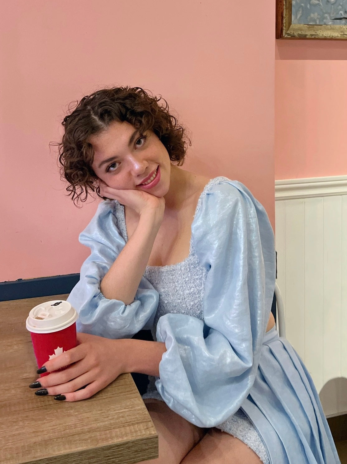a model with short curly brown hair smiling at the camera holding a coffee cup and wearing a baby blue bustier and skirt set