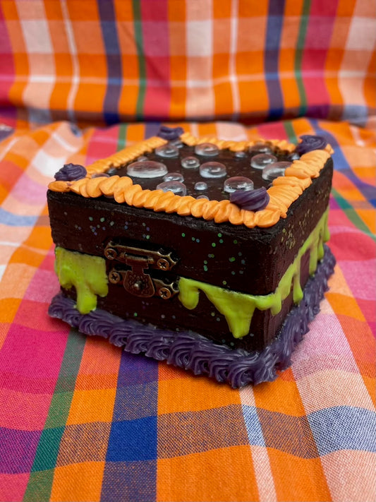 a small hinged box painted black and decorated to look like a cake with black, purple, and orange icing and with green drips on the side