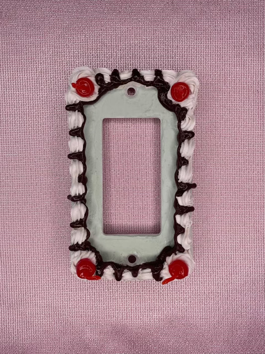 a light switch plate decorated to look like a mint green cake with fake cherries and chocolate drizzle