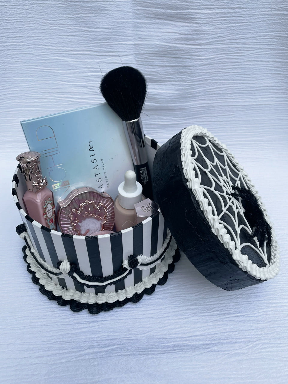 an opened black and white cardboard box decorated to look like a cake and holding various makeup items