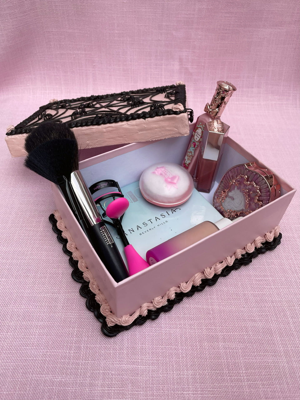 an open cardboard box decorated like a pink and black cake and holding various makeup items