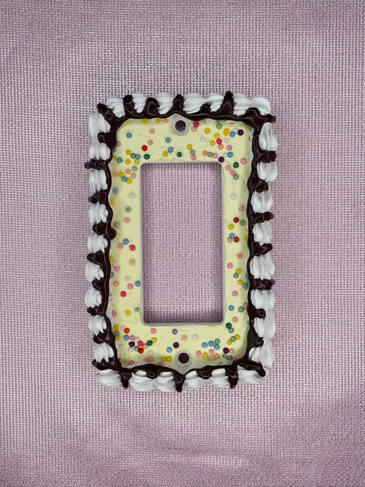a light switch plate decorated to look like a vanilla cake with sprinkles and chocolate drizzle